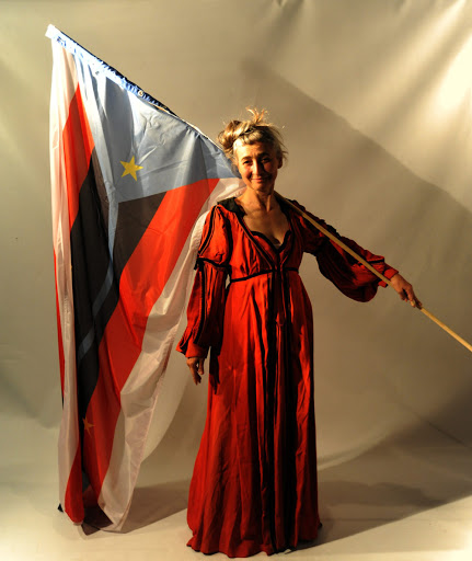 Starsza kobieta ubrana w czerwoną suknię, trzymająca flagę