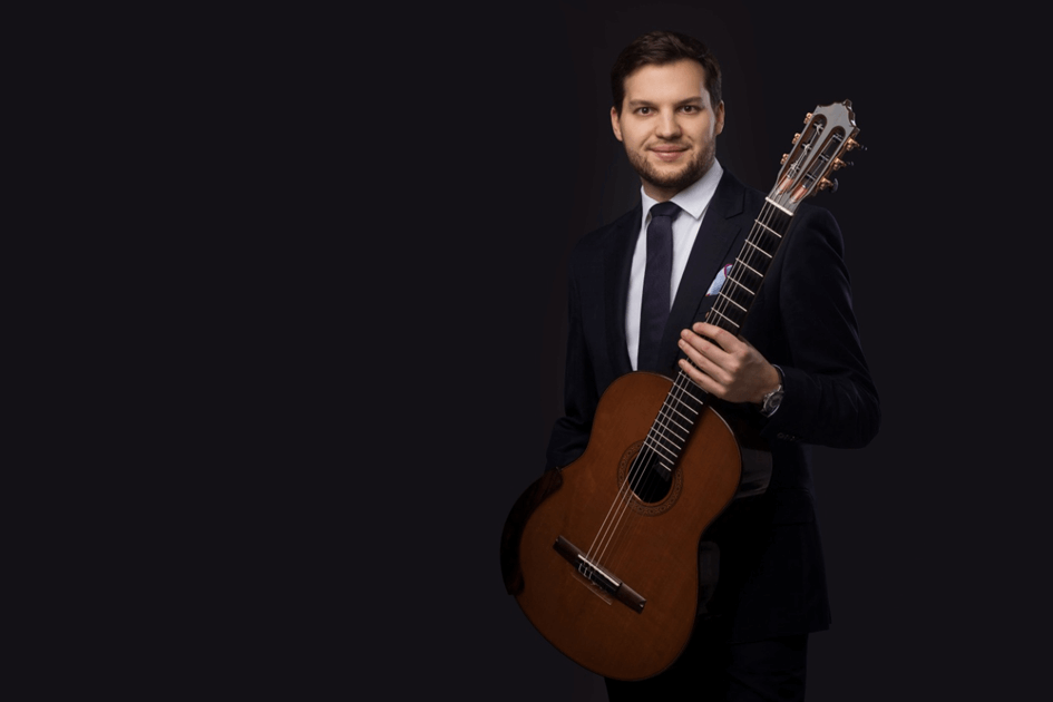 Jakub Kościuszko pozuje do zdjęcia w garniturze z gitarą w ręku