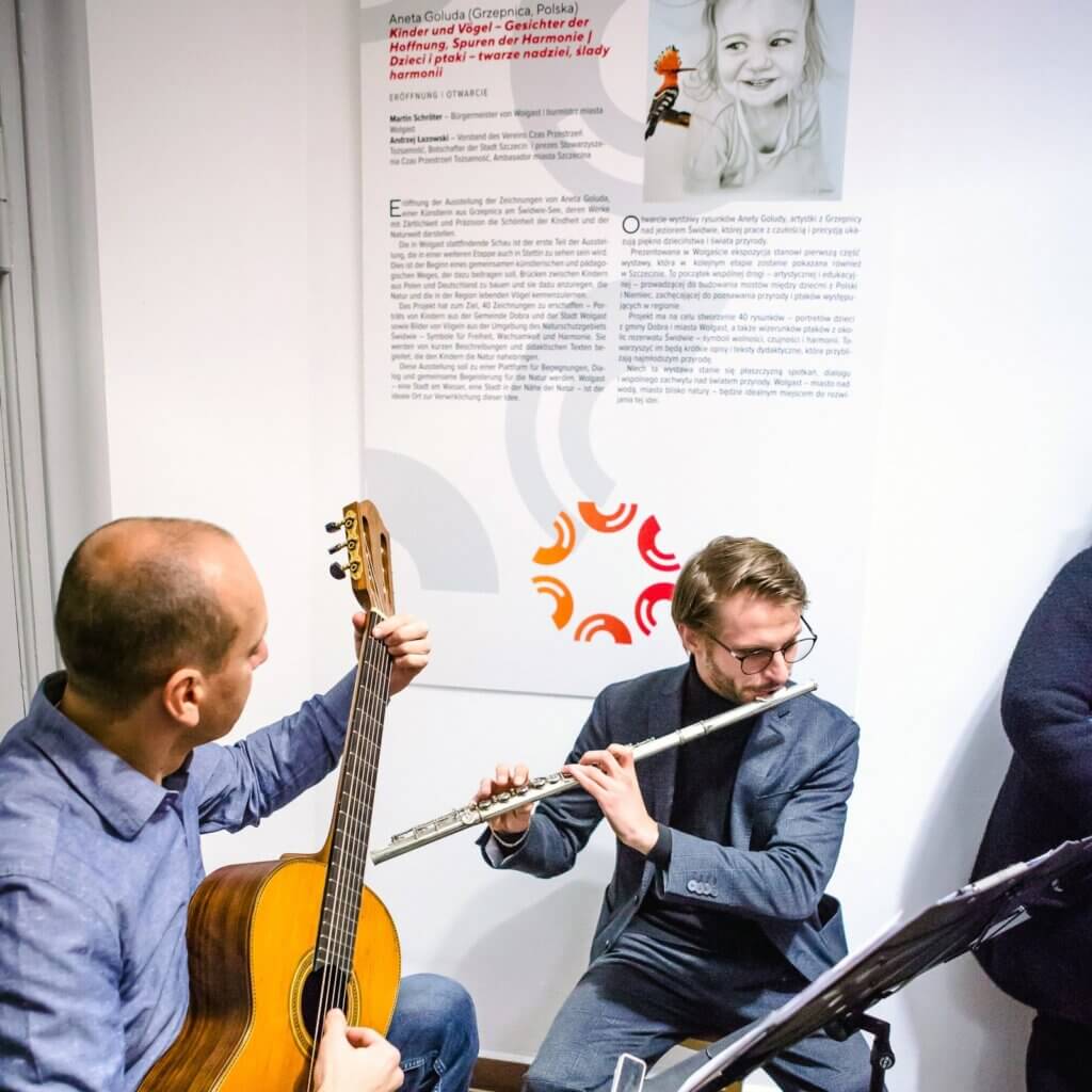 Gitarzysta i flecista grają podczas otwarcia wystawy w Wolgaście, w tle plansza wystawy